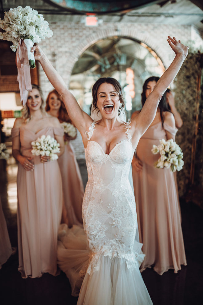Bride in couture wedding dress with arms raised and bridesmaids with blush dresses holding petite bouquets stand behind her inside the grand hall at the Astorian