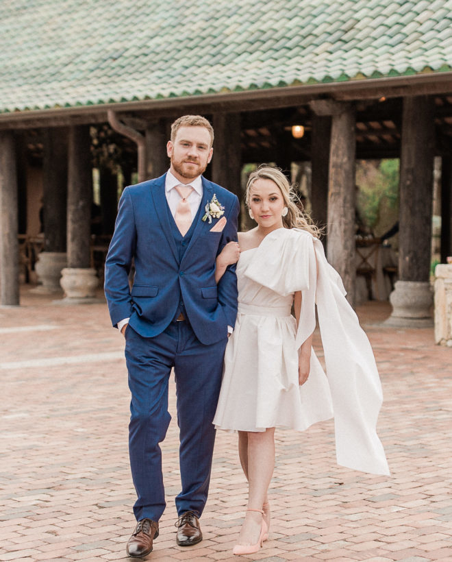 Dramatic shot of bride, in reception mini dress with statement bow and heels, and groom, in navy suit with blush accents, looking directly at camera outside of the Camp Lucy outdoor pavilion
