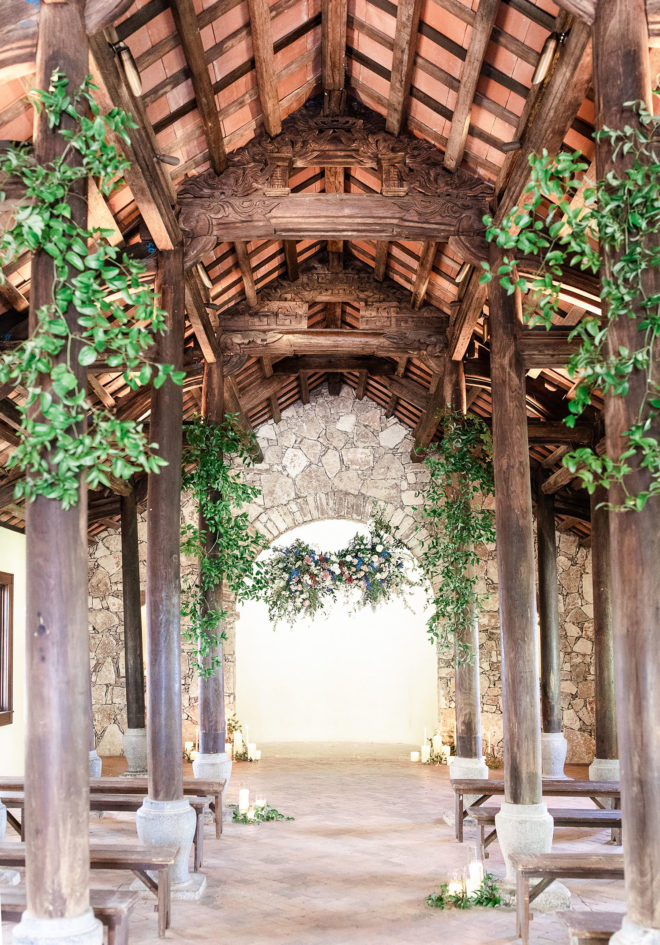 Interior of Ian's Chapel at Camp Lucy unfinished wood rafters and posts accented with with fresh greenery and lush blush, wine organic florals and lit candles encased in clear glass candleholders with greenery wrapped around