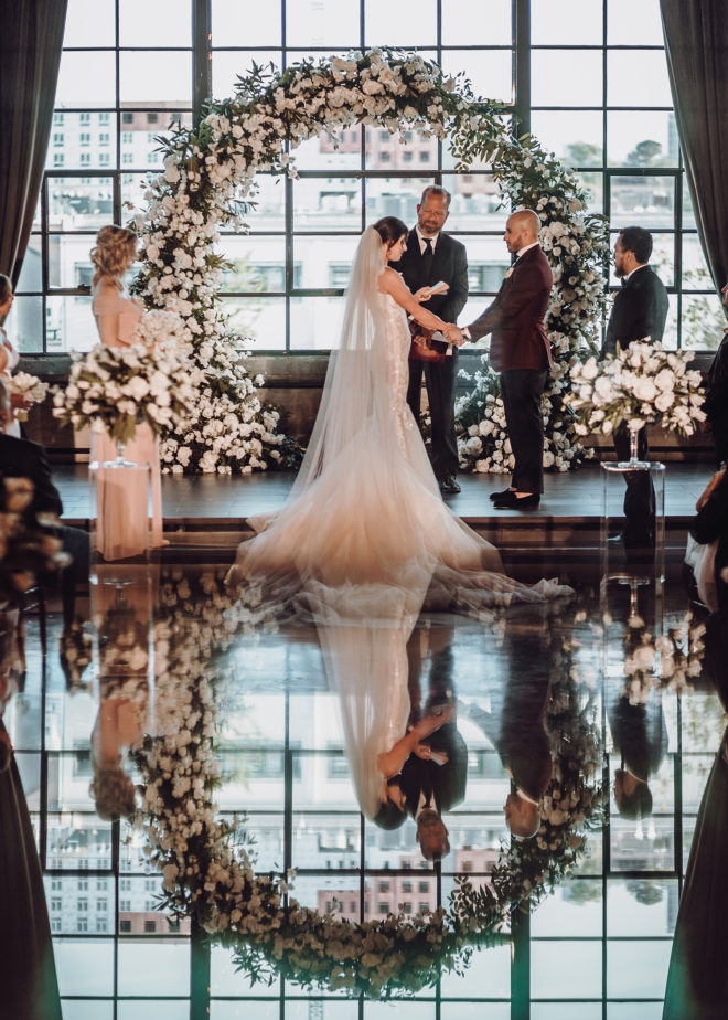 Bride and groom holding hands at end of aisle saying vows with Ecclesia Church Pastor Chris Seay with large flower arch and large loft window overlooking Houston at the Astorian