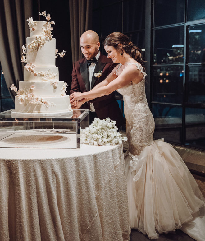 Bride and groom cutting 5 tiered cream colored cake with delicate florals, brides couture dress is bustled and both bride and groom are smiling happily in the industrial chic indoor ballroom