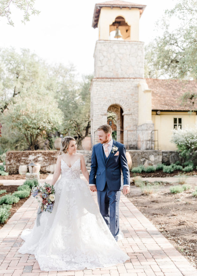 Bride, in lace gown, holding pastel bouquet and groom holding hands on sunny day with exterior of Ians Chapel at Camp Lucy behind the couple
