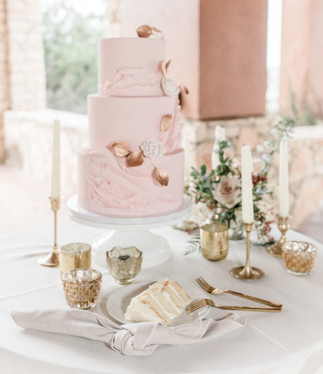 White linen cake table underneath al fresco pavilion with three tiered blush colored cake adorned with rose gold gilded fondant leaves and fondant florals surrounded by vintage style mercury glass votives and tall white tapered candles held by gold candlesticks 