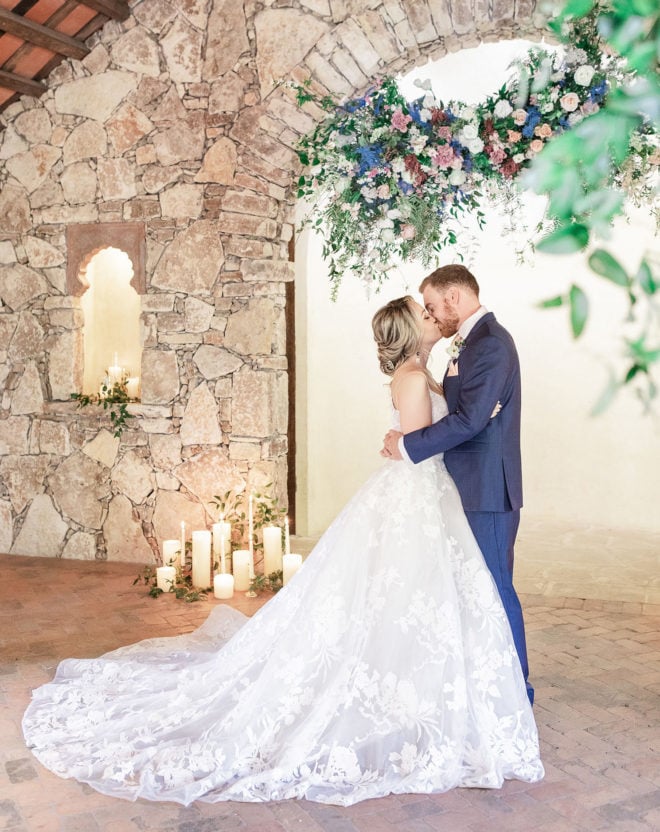 Bride and groom kissing inside Camp Lucys Ian Chapel surrounded by greenery, a pastel colored hanging floral installation and glowing candles 