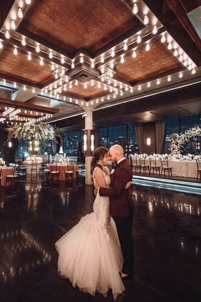 Bride and groom standing in center of Astorians industrial chic grand hall embracing each other as if slow dancing in the empty room lit up vintage new york style ceiling lighting and grand floral centerpieces surrounded by candlelight