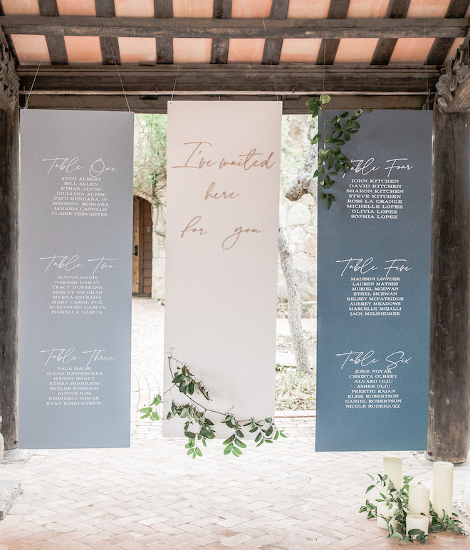Hanging installation of large custom panels with seating assignments in cursive and middle panel which reads, "Ive waited here for you" with greenery attached