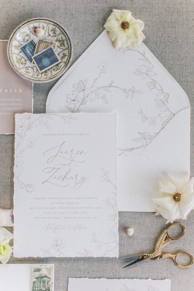 Flay lay photo of white letterpress wedding invitation suite with delicate flowers, vintage stamps, and two scattered white flowers 