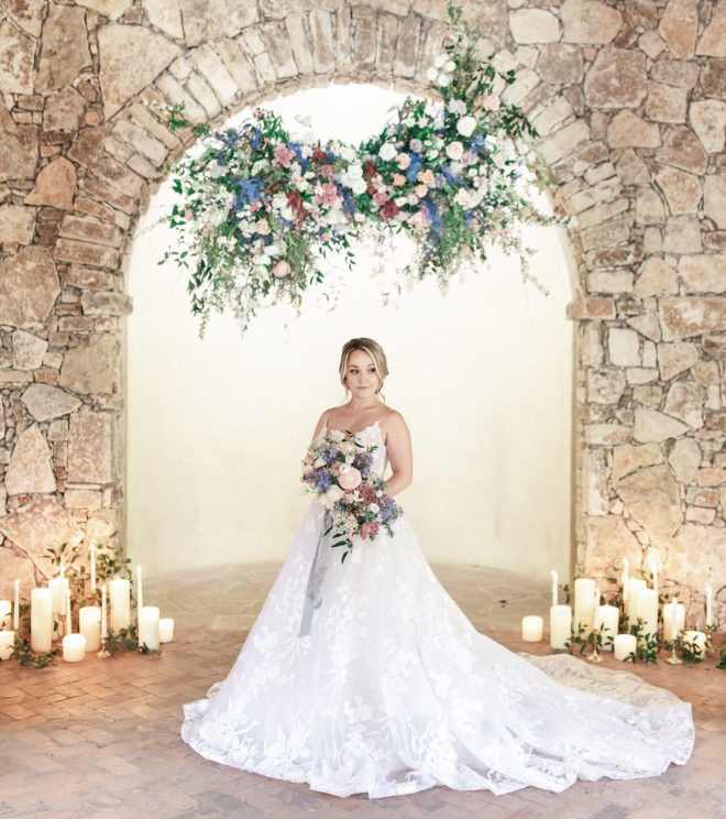 Bride with long traditional gown holding pastel colored bouquet of florals surrounded by white block and pillar candles and greenery inside of Camp Lucy Ians Chapel 