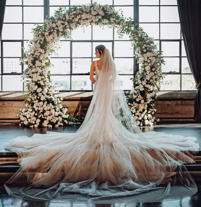 Bride, with long flowing veil, stands in front of blooming floral arch and large sunny loft window before the wedding reception at the Astorian 