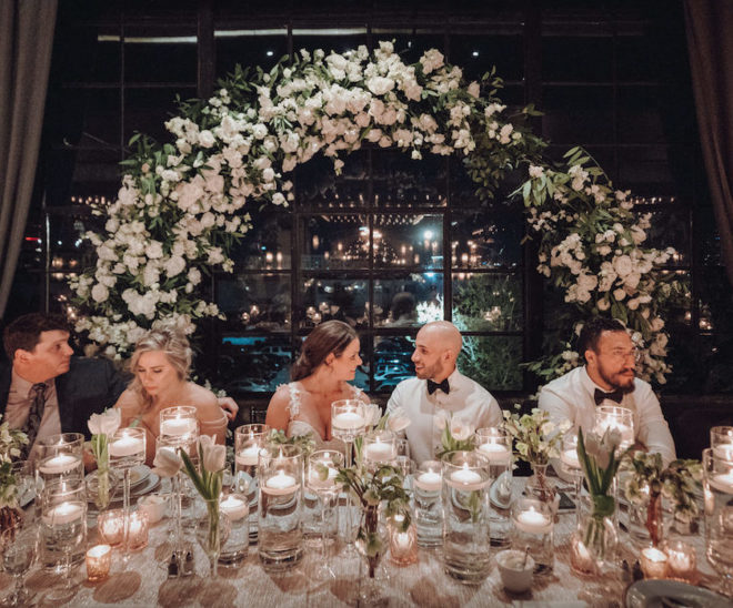 Cream colored party tablescape with glass cylinders filled with water and floating votives, fresh cut tulips and large statement backdrop with white florals, bride and groom are smiling at each other while seated at the wedding table