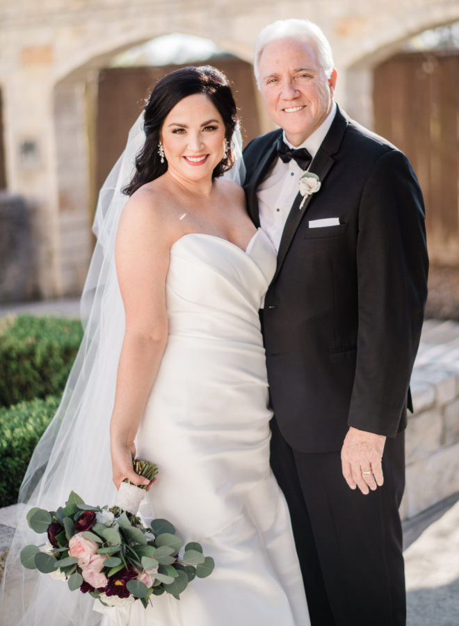 Bride with bouquet of blush and wine colored flowers standing with her father in a black suit outside by green bushes and white austin stone facade of briscoe manor wedding venue