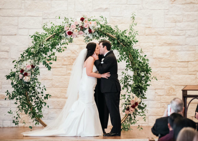 Couple kissing in front of arched alter of blush and wine florals. 