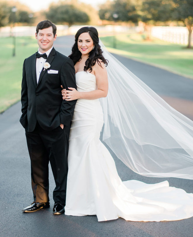Groom in black tuxedo and bride in white flowing gown and veil standing outside on paved road at venue, Briscoe Manor