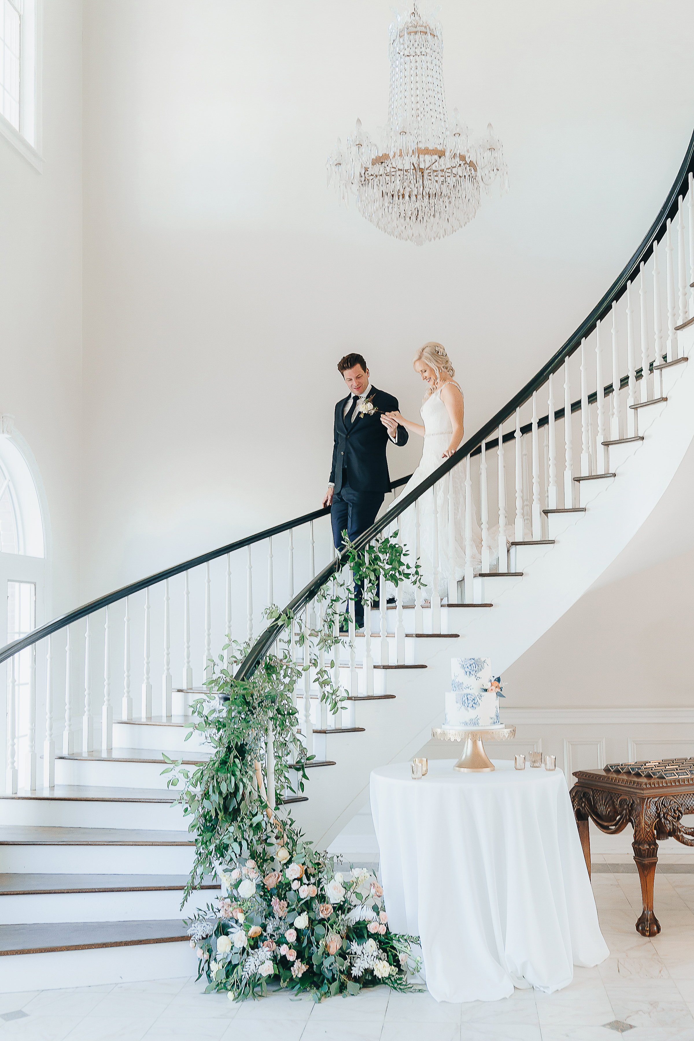 Groom walking the Bride down a curving staircase with their cake display at the bottom