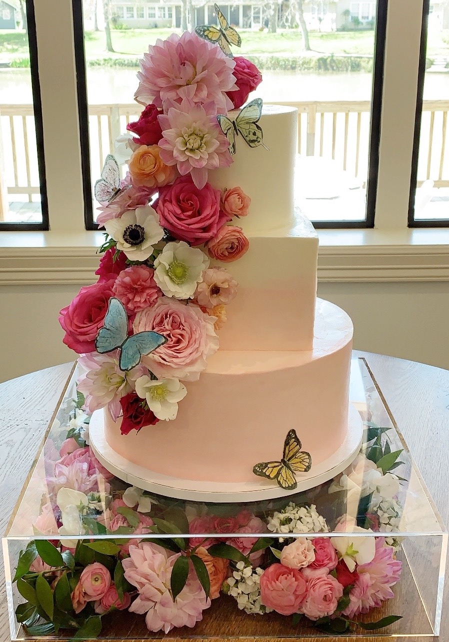 three tier pink to white ombre cake with pink florals and paper butterflies. On acrylic cake stand filled with flowers.
