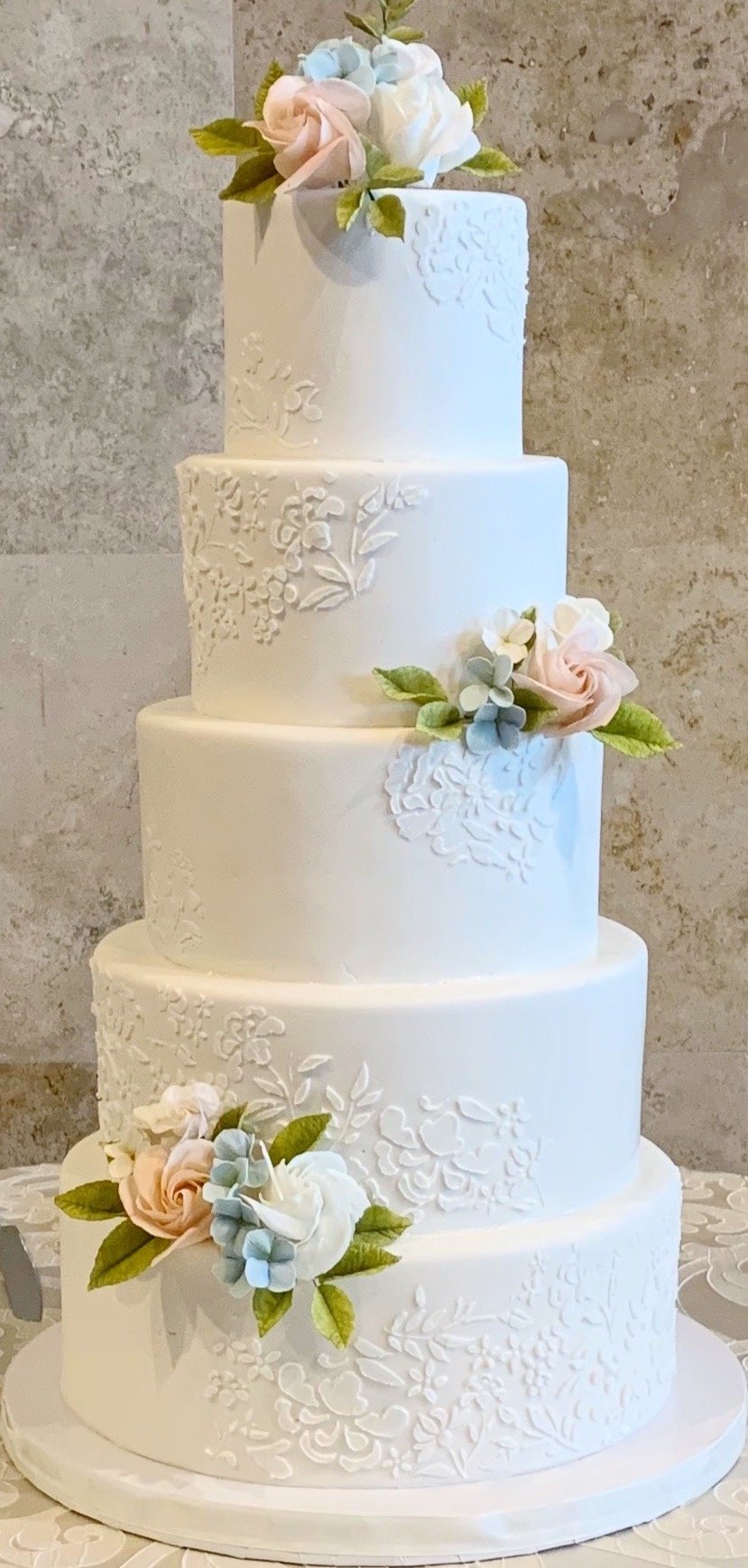 White 5 tier cake with white flower stencil design and light pink and blue 3D flowers.