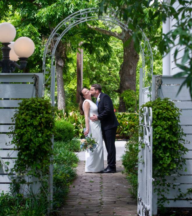 Groom, in black suit, kissing the cheek of bride, in long white simple gown, in the lush green gardens of Heather's Glen wedding venue in Conroe, Texas.