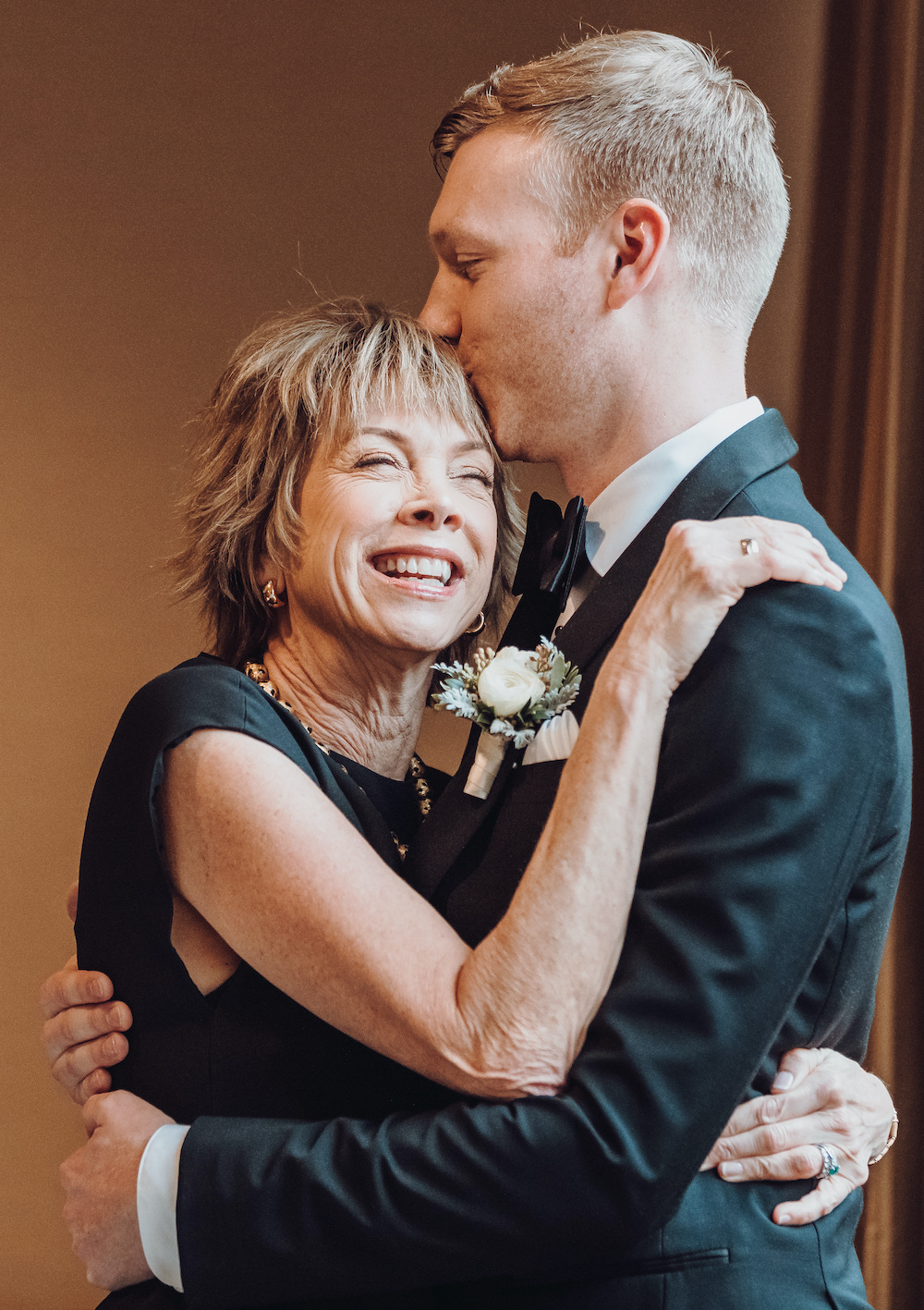 Groom in black tuxedo embracing his mother as they laugh. 