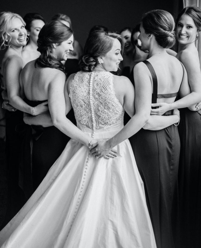 Black and white photograph of bride, wearing long white gown with lace back, hugging group of bridesmaids in various updos wearing sleeveless black silky gowns.