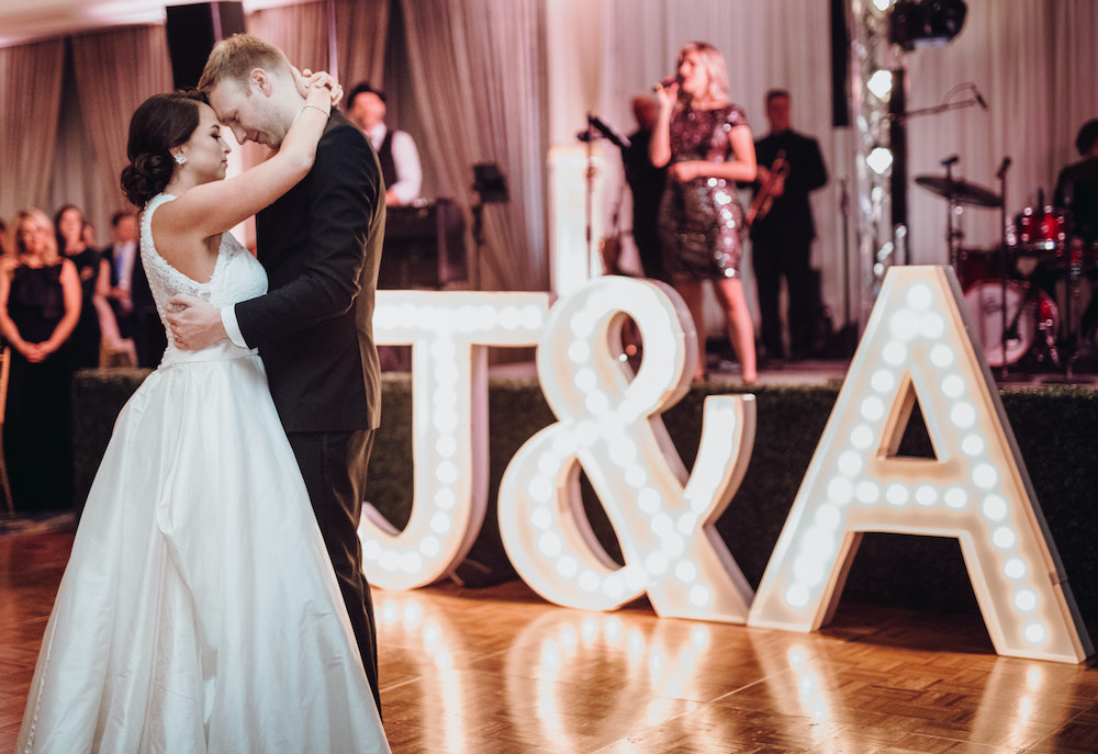 Bride, in white gown with lace back and low updo, dancing with groom, in all black tuxedo, on light wood dance floor with tall lit marquee letters which reads, "J&A" 