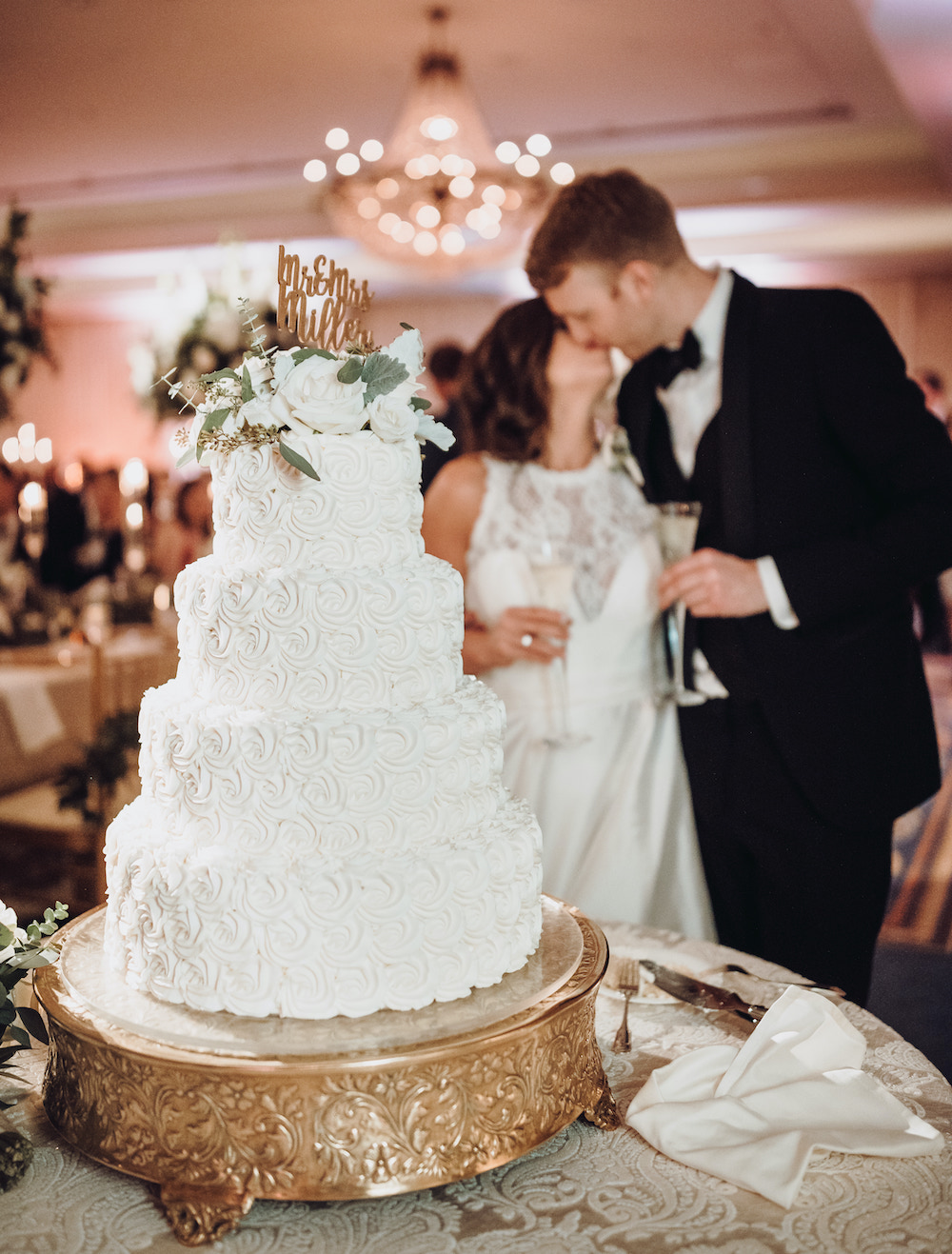 White four tier cake with white blooms and custom lettering as topper which reads, "Mr. and Mrs. Miller" and bride and groom in background kissing and holding champagne flutes 