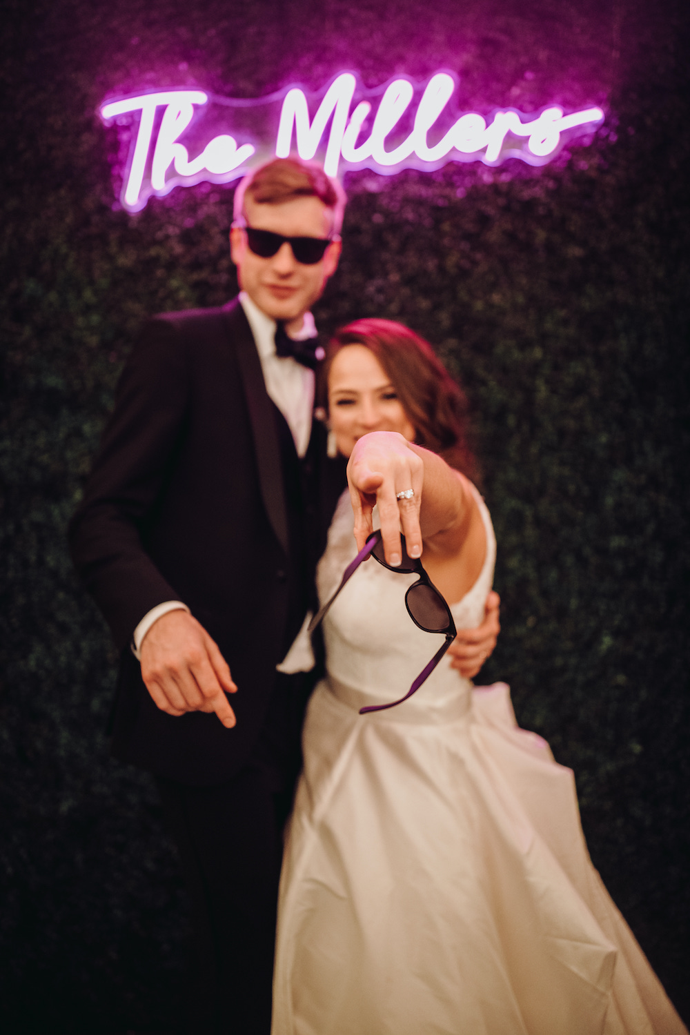 Groom, wearing sunglasses and bride pointing to camera with sun glasses in hand, in front of greenery statement wall with fuchsia colored LED sign that reads, "The Millers"