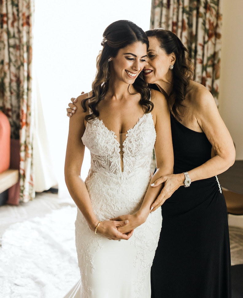 Mother of the bride in a black, one shoulder gown hugging the bride to be wearing a gorgeous lace wedding gown. 