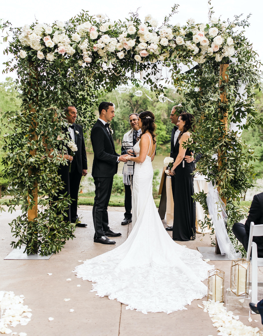 Bride and groom saying their vows under a chuppah beautifully decorated with greenery and topped with an assortment of flowers in blush and white.