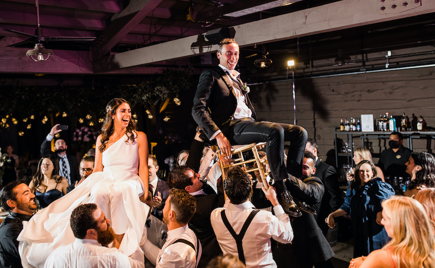 The bride and groom are seated on chairs and lifted in the air during the celebratory dance at the reception called the hora. where guests dance in a circle. 