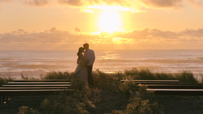 The bride and groom stand at the end of the wedding aisle with the sun setting over the Pacific ocean in the background after this Oregon destination wedding.