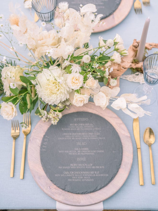 The wedding table setting for this Oregon Coast destination wedding includes wooden chargers, an engraved slate menu, gold flatware, and white flowers.