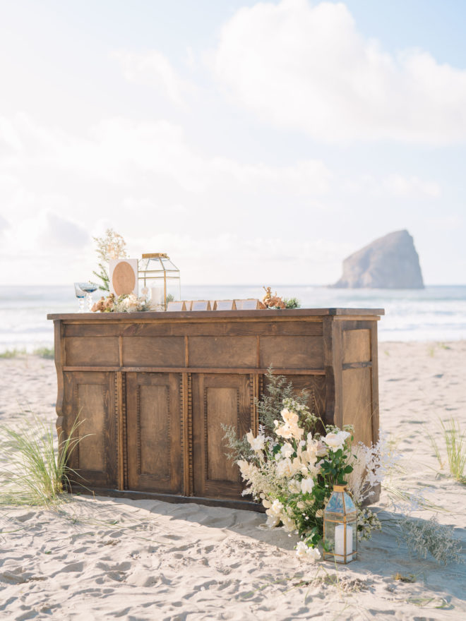 Rustic, wooden wedding escort table with white wedding flowers, gold accents, and Haystack Rock in the background.