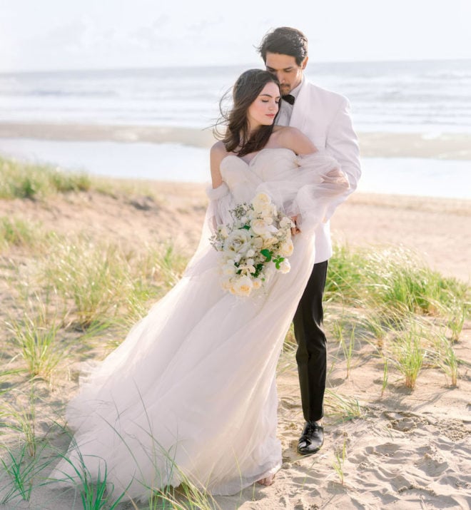 Bride, wearing off the shoulder white tulle gown holding white monochromatic bouquet, held from behind by a groom wearing a white tuxedo shirt, black bowtie and black pants on Oregon's Cannon Beach.