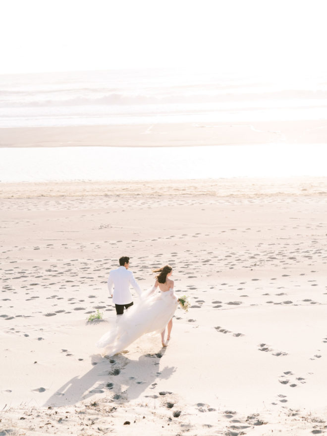 Ariel shot of Groom in white tuxedo jacket and black pants holding hands with bride in flowing white tulle gown on sun dappled Oregon Cannon beach