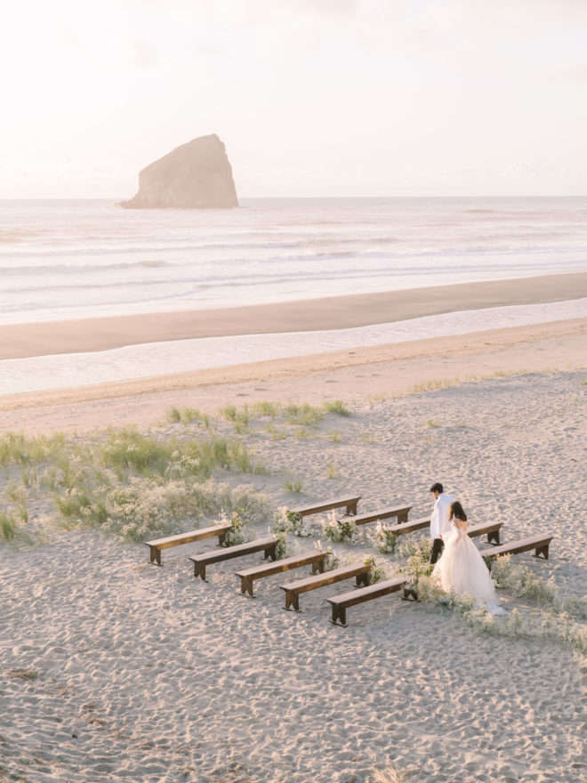 A wedding aisle is created on Cannon Beach with white wedding floral arrangements on each side separating the backless wooden benches.
