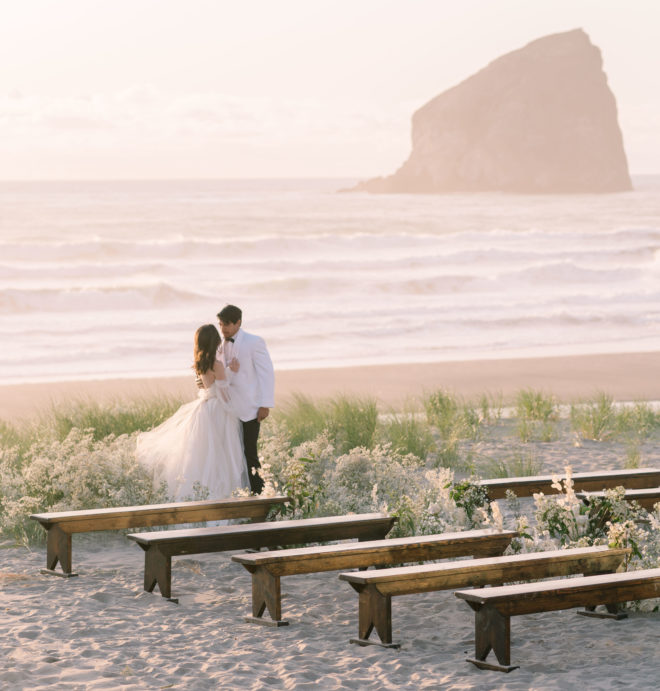 Bride and groom standing at end of al fresco wedding aisle amidst tall green beach botanicals with crashing waves and large rock formation behind them on Oregons Canon Beach