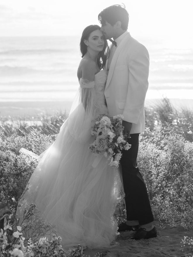 Black and white photo of bride, wearing tulle off the shoulder flowing gown holding bouquet of florals, looking directly at camera while groom, wearing white tuxedo jacket and black pants, kisses her forehead amidst beach botanicals on Cannon Beach.