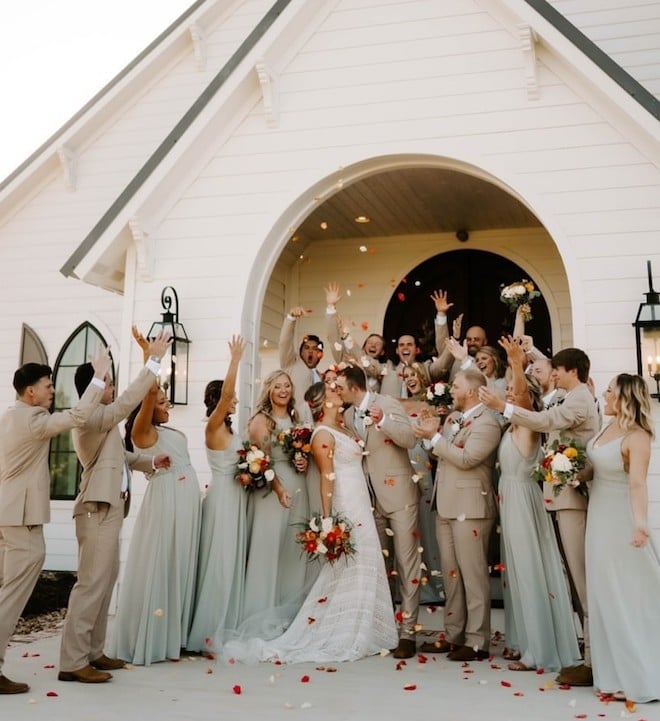 Bridesmaides and Groomsmen in sage and tan throwing flower petals around a bride and groom kissing. 