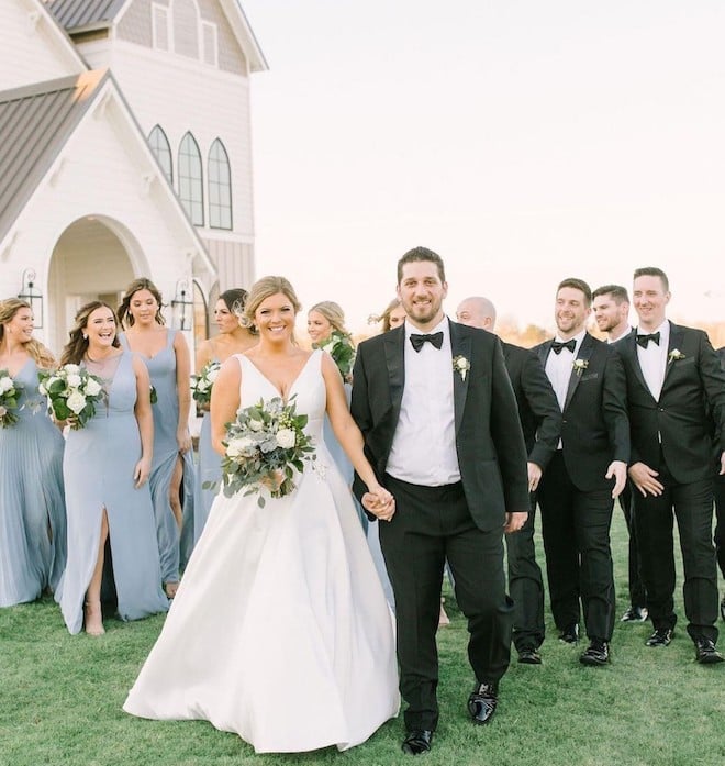 Bride and groom walking in front of bridesmaids in blue and groomsmen in black. 