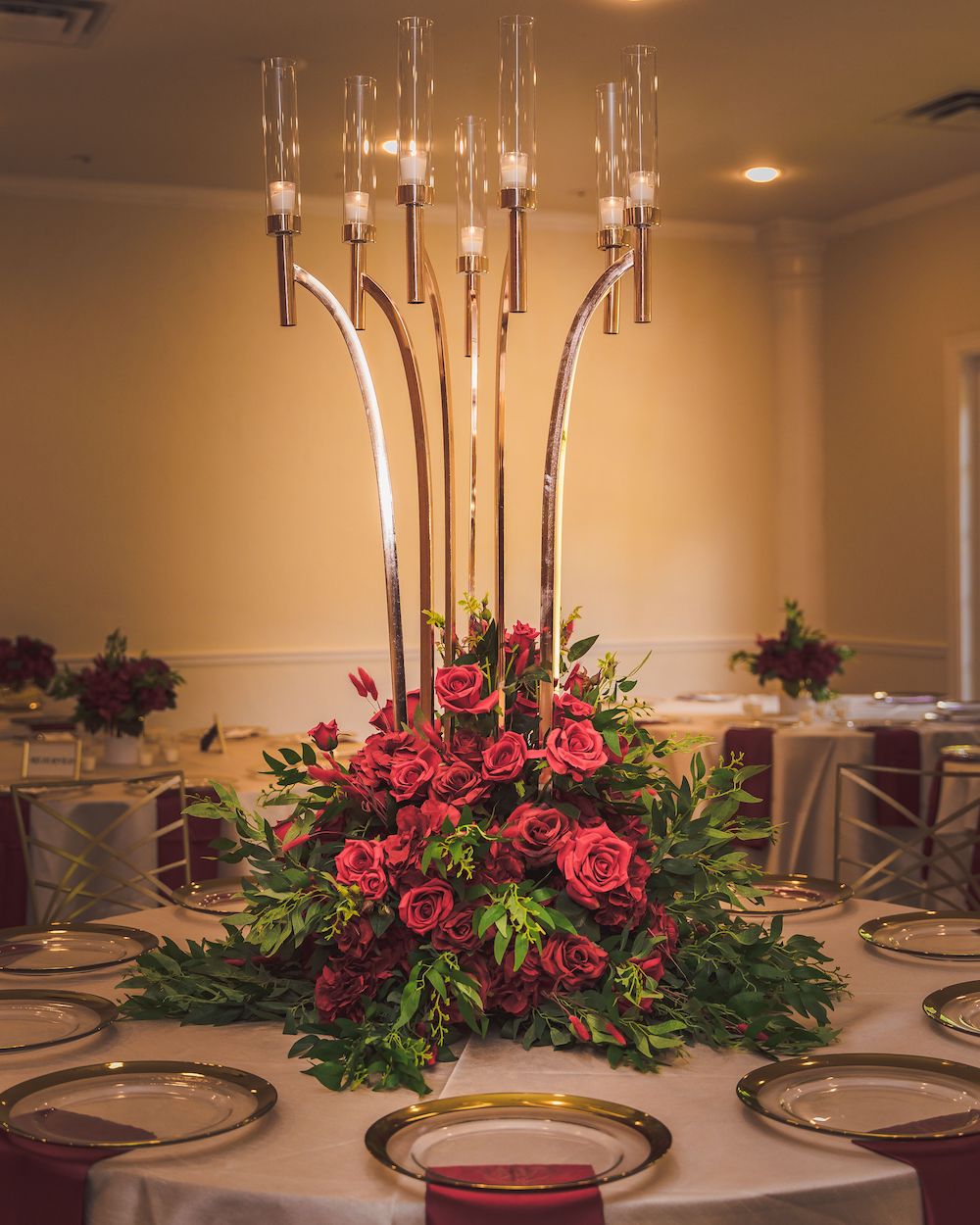 Tall gold and glass candle holders above a centerpiece arrangement of red roses.