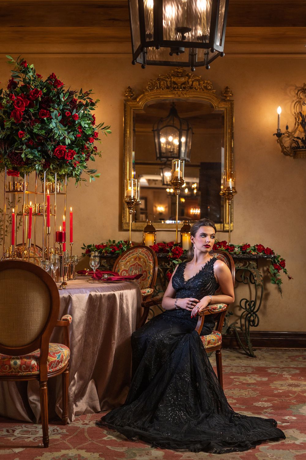 Bride with red lips, black smokey eye makeup and ponytail in sleeveless black wedding gown seated at table with tall blooming red rose centerpiece, red pillar candles with gold candleholders, and metallic table skirt inside of ballroom of Hotel Granduca Houston.