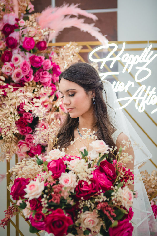 Bride, holding hot pink, blush and ivory bouquet of roses, with hair tied back with veil, amongst hot pink, gold and blush plumes adhered to a gold backdrop with an LED sign that reads, "Happily Ever After" in the ballroom of Hyatt Regency Hotel Houston. 