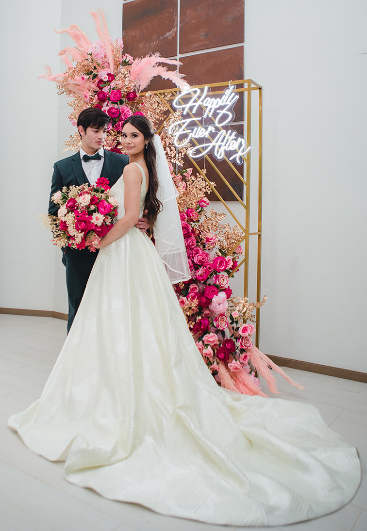 The bride and groom stand in front of a city chic photo backdrop adorned with fuchsia, blush, and gold florals.