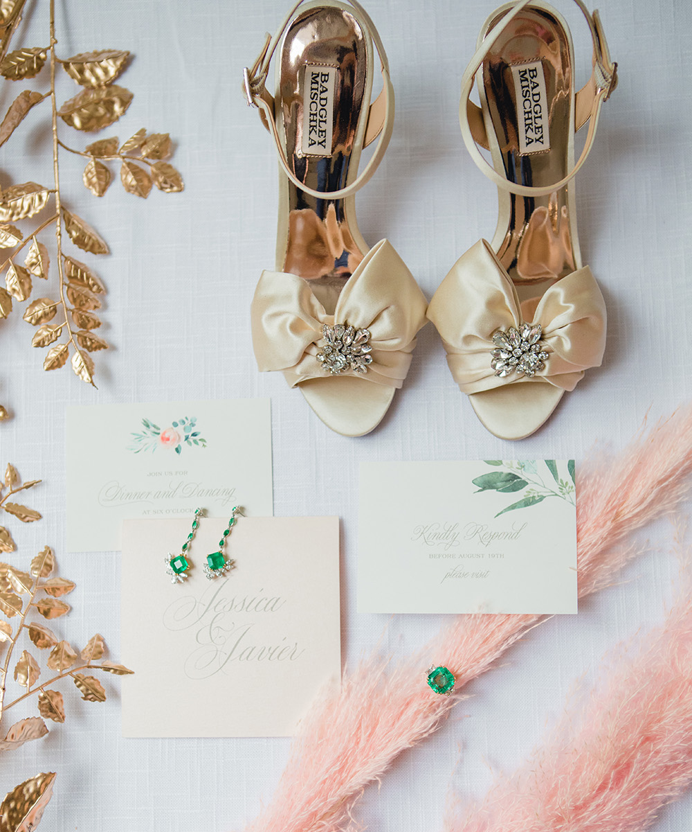 Wedding invitations rest atop white canvas surrounded by gold and blush foliage, gold heels, and emerald jewelry.