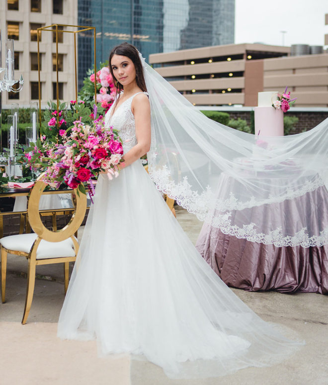 Bride, on the outside garden and patio of Hyatt Regency Hotel Houston, holding blush and hot pink bouquet of florals, in long white tulle gown with long formal veil flowing directly behind her.