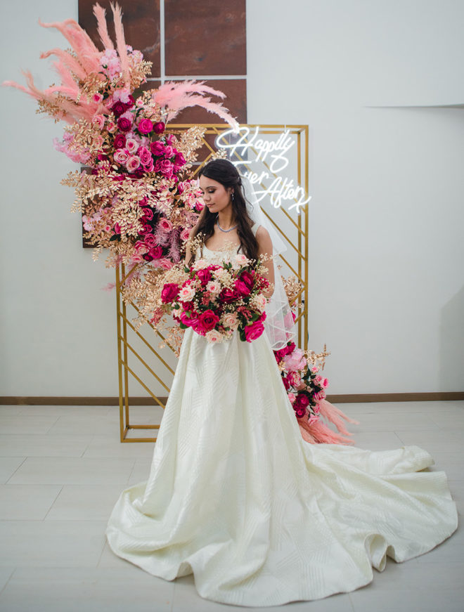 Bride holding bouquet of hot pink and blush florals with gold backdrop accented with lush blush colored plumes, coordinating hot pink and blush florals, gold leaves and LED sign which reads, "Happily Ever After"