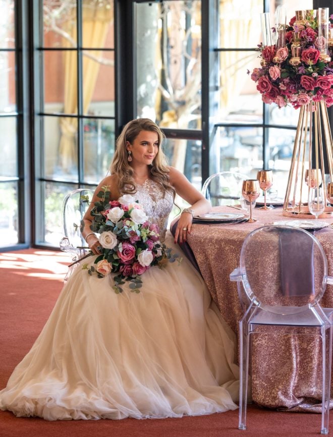 Bride, in tulle skirted ball gown holding bouquet of silk ivory, peach and mauve colored roses, sitting at reception table with dusty pink table skirt, rose gold glasses and 
