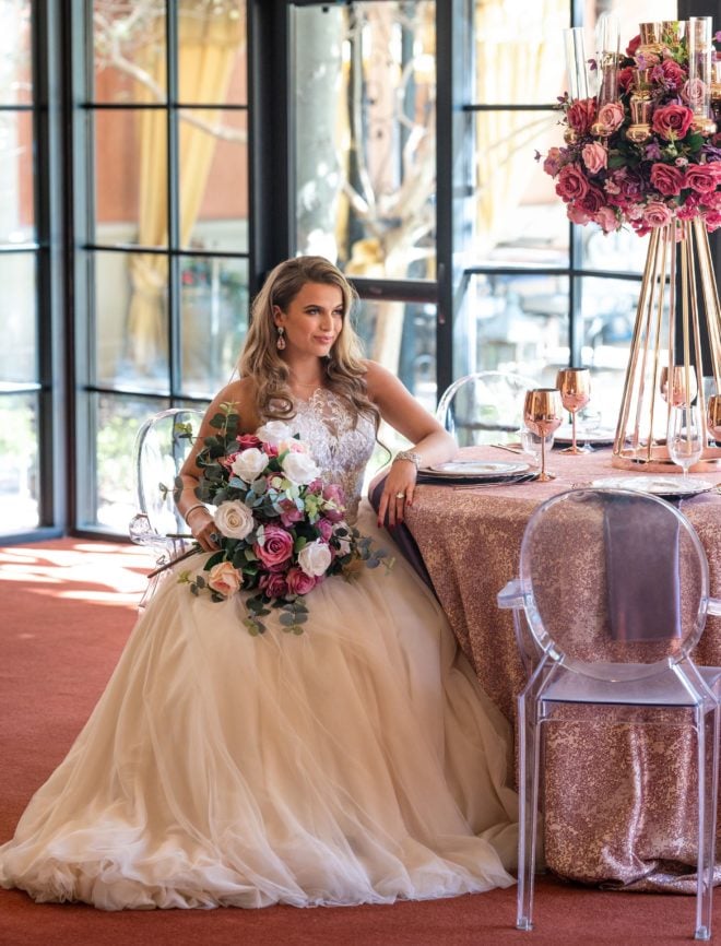 Bride, in tulle skirted ball gown holding bouquet of silk ivory, peach and mauve colored roses, sitting at reception table with dusty pink table skirt, rose gold glasses and