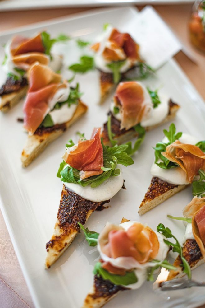 Flatbread sandwich bites topped with fig puree mozzarella, aged prosciutto di Parma, and arugula.