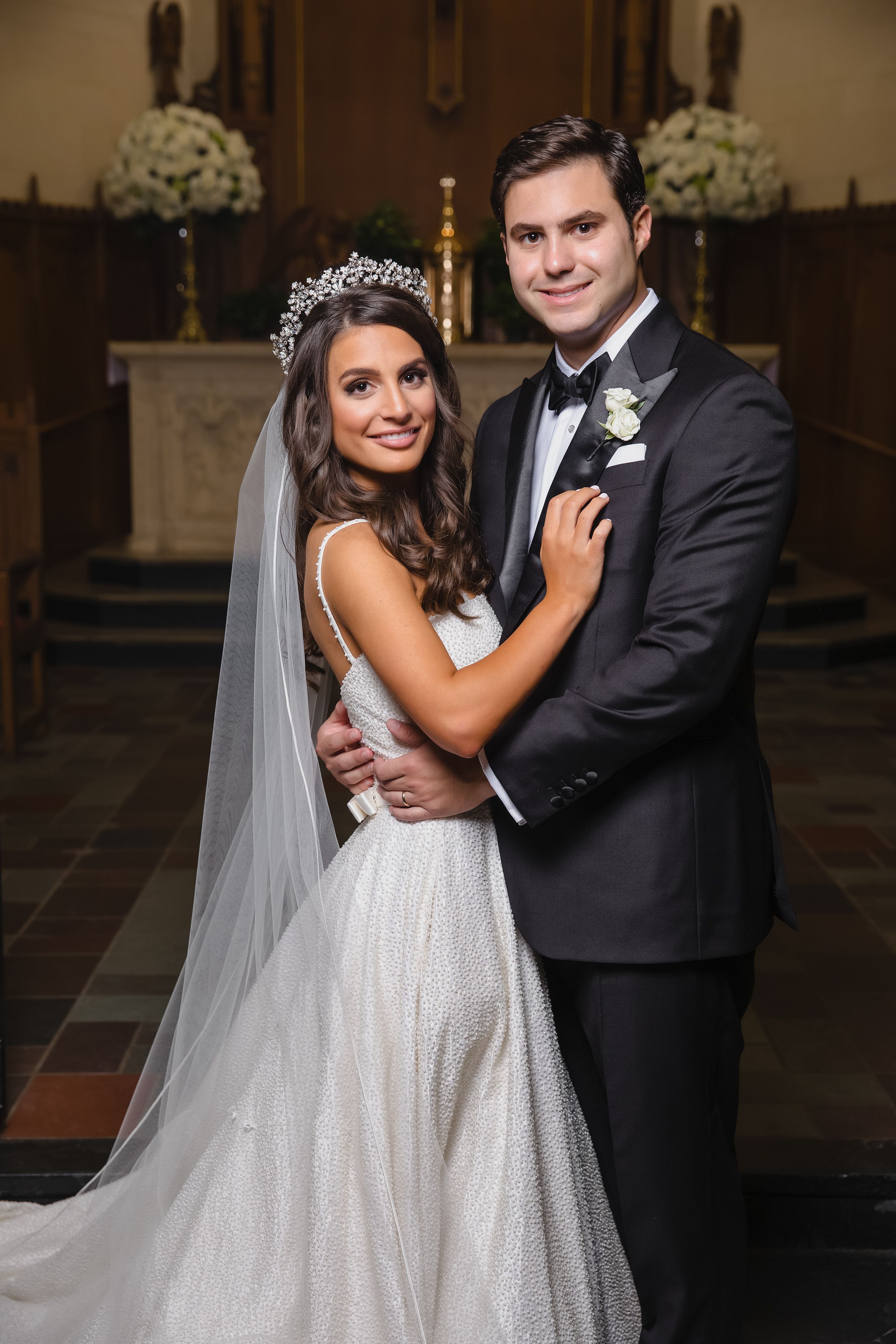 A bride, with ornate headpiece and long veil and white textured ball gown, standing groom, in all black tuxedo,  after getting married in Houston at a Catholic church.